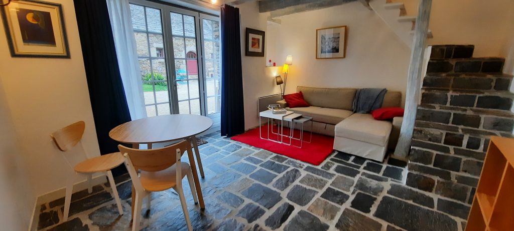 Pièce de vie du gîte Anita Conti avec canapé d'angle, table ronde et sol en dalles de pierre